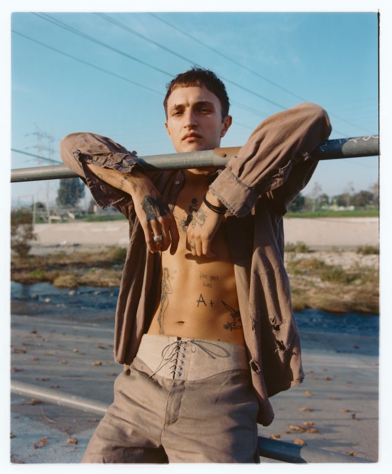 Anwar Hadid wearing an open taupe shirt and lace-up trousers leaning over a metal railing outdoors.