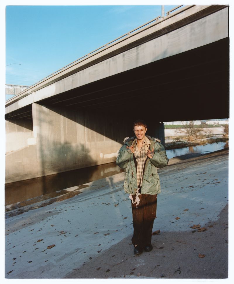 Model Anwar Hadid smiling under a concrete highway bridge wearing an oversized green parka jacket and brown trousers.