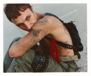 Anwar Hadid seated outdoors wearing green cargo trousers and a black strap bag, showcasing numerous tattoos.