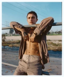 Anwar Hadid wearing an open taupe shirt and lace-up trousers leaning over a metal railing outdoors.