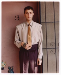 Anwar Hadid standing in a doorway wearing a beige collared shirt, gold silk tie, and high-waisted dark trousers.