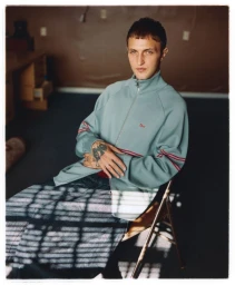 Anwar Hadid seated on a folding chair wearing a light blue zip-up jacket and dark trousers for Behind the Blinds.