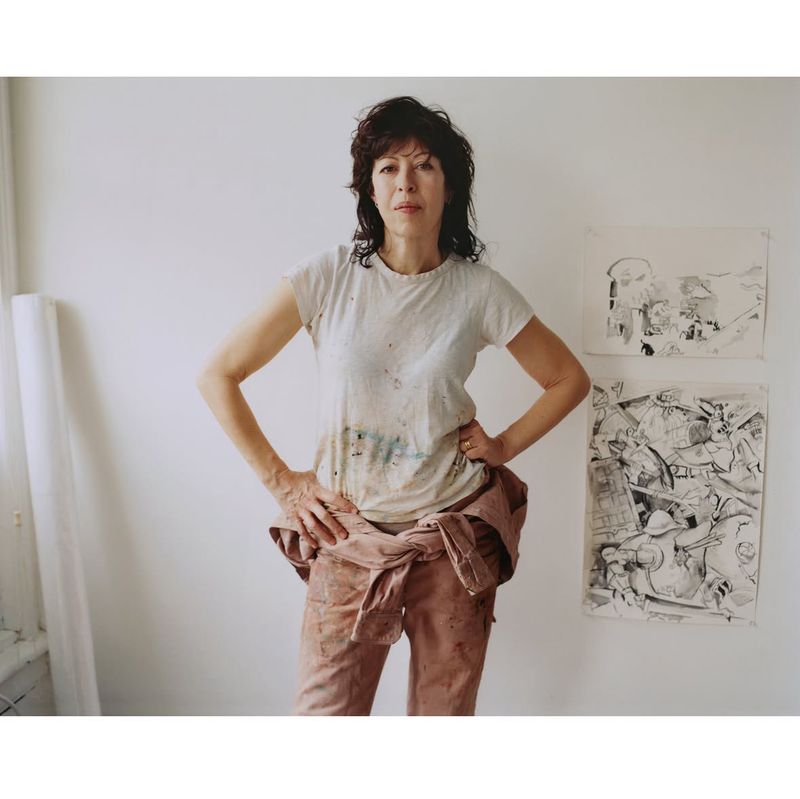 Portrait of a woman wearing a white t-shirt and pink trousers standing against a plain white wall with sketches.