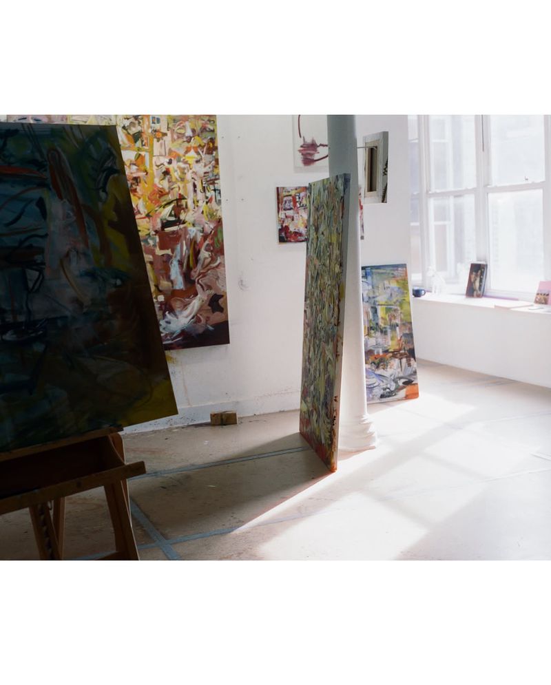 Sunlit artist studio interior with abstract oil paintings on easels and resting against walls and a central pillar.
