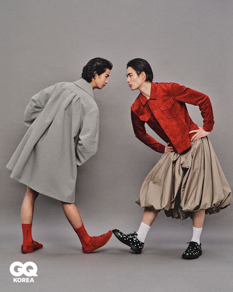 Two male models facing each other against a grey studio backdrop, wearing styled coats, skirts, and patterned socks.