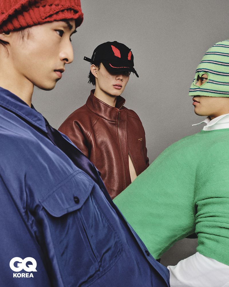 Three male models wearing colorful streetwear including a blue jacket and green balaclava against a grey studio backdrop