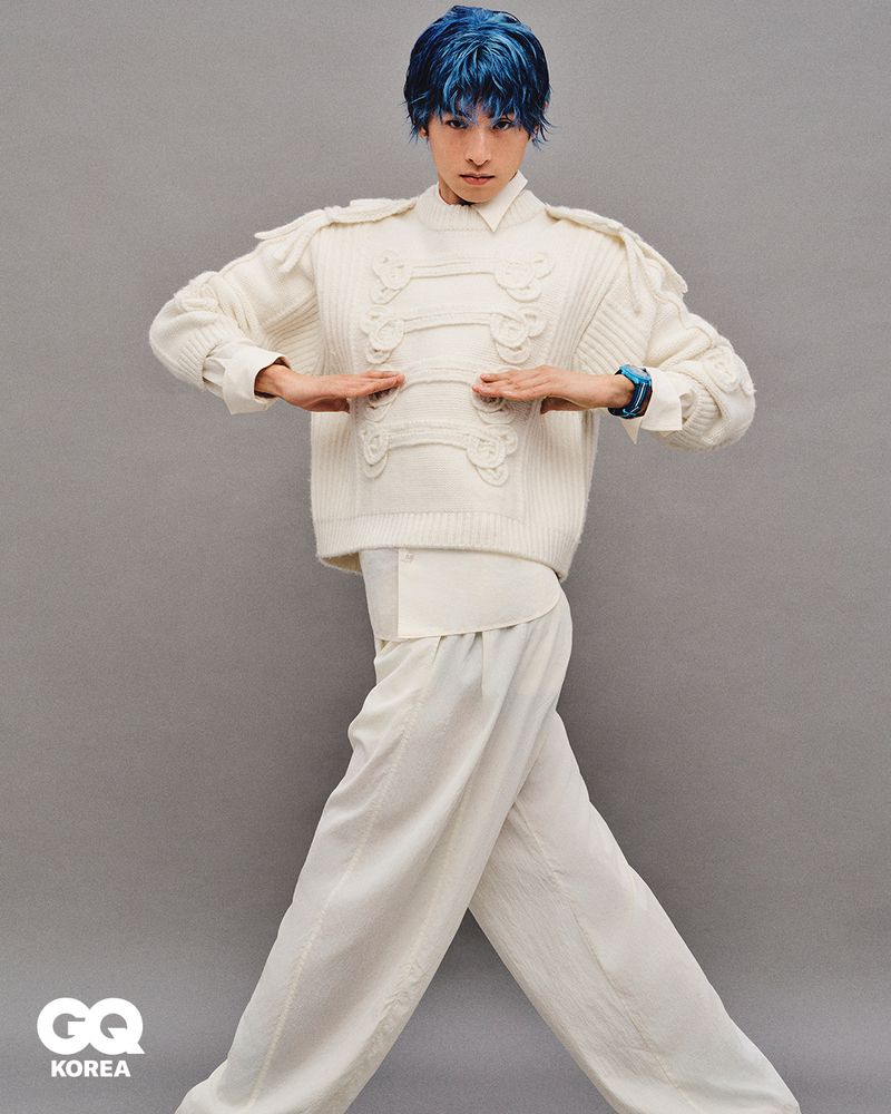 Model Yugo Takano with blue hair wearing a cream military-style knit sweater and wide-leg trousers against a grey backdrop for GQ Korea.