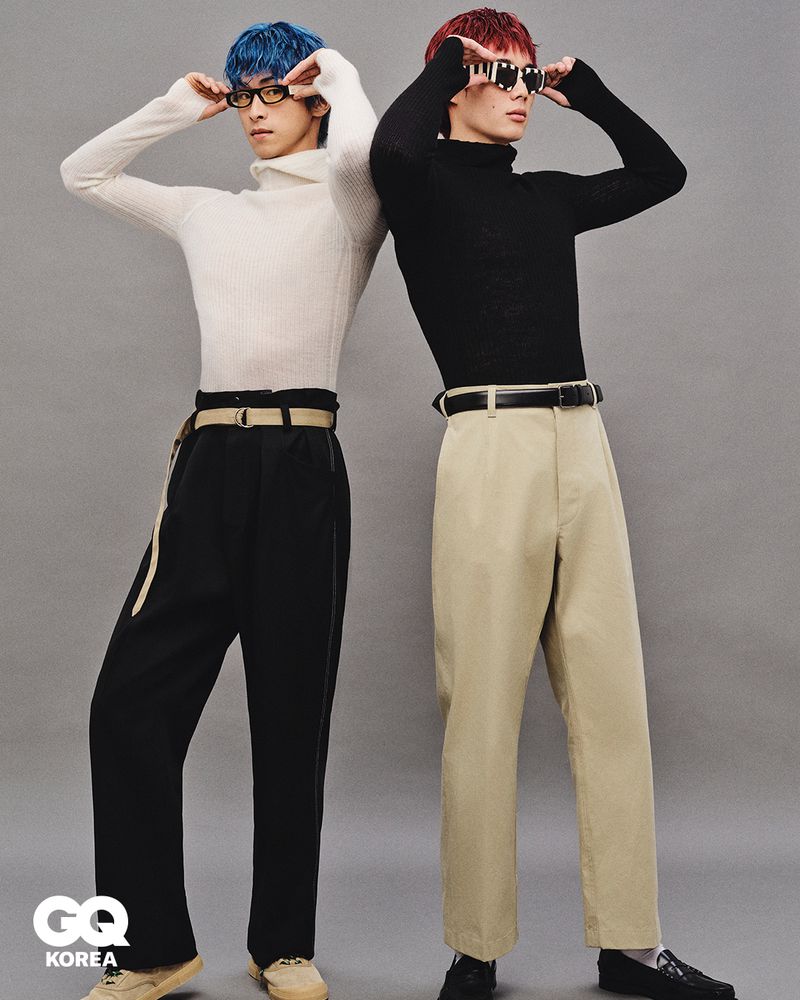 Two men pose back-to-back indoors against a grey wall. They wear colorful hair, turtlenecks, trousers, belts, and sunglasses.