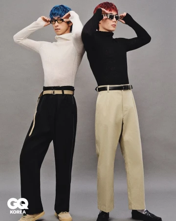 Two men pose back-to-back indoors against a grey wall. They wear colorful hair, turtlenecks, trousers, belts, and sunglasses.