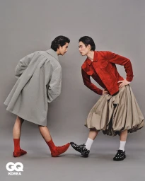 Two male models facing each other against a grey studio backdrop, wearing styled coats, skirts, and patterned socks.