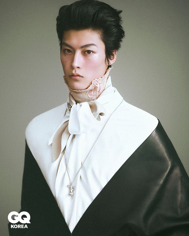 Portrait of a male model wearing a white cravat shirt and black jacket against a neutral grey studio background