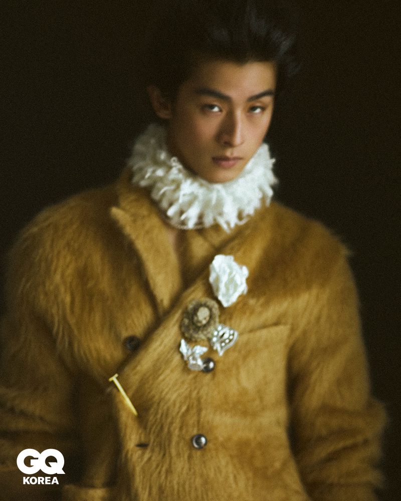 Model Yugo Takano wearing a textured camel hair coat and white pleated neck ruff against a dark background for GQ Korea.