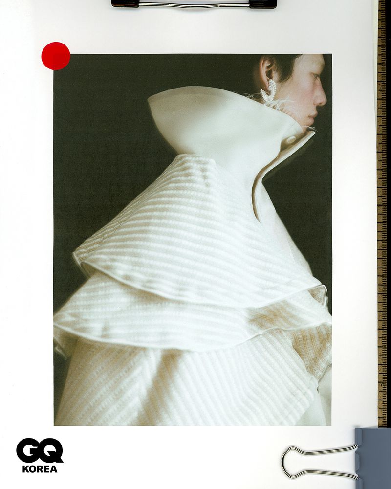 Model wearing a structured white high-collar ruffled garment in profile view against a plain background for GQ Korea.