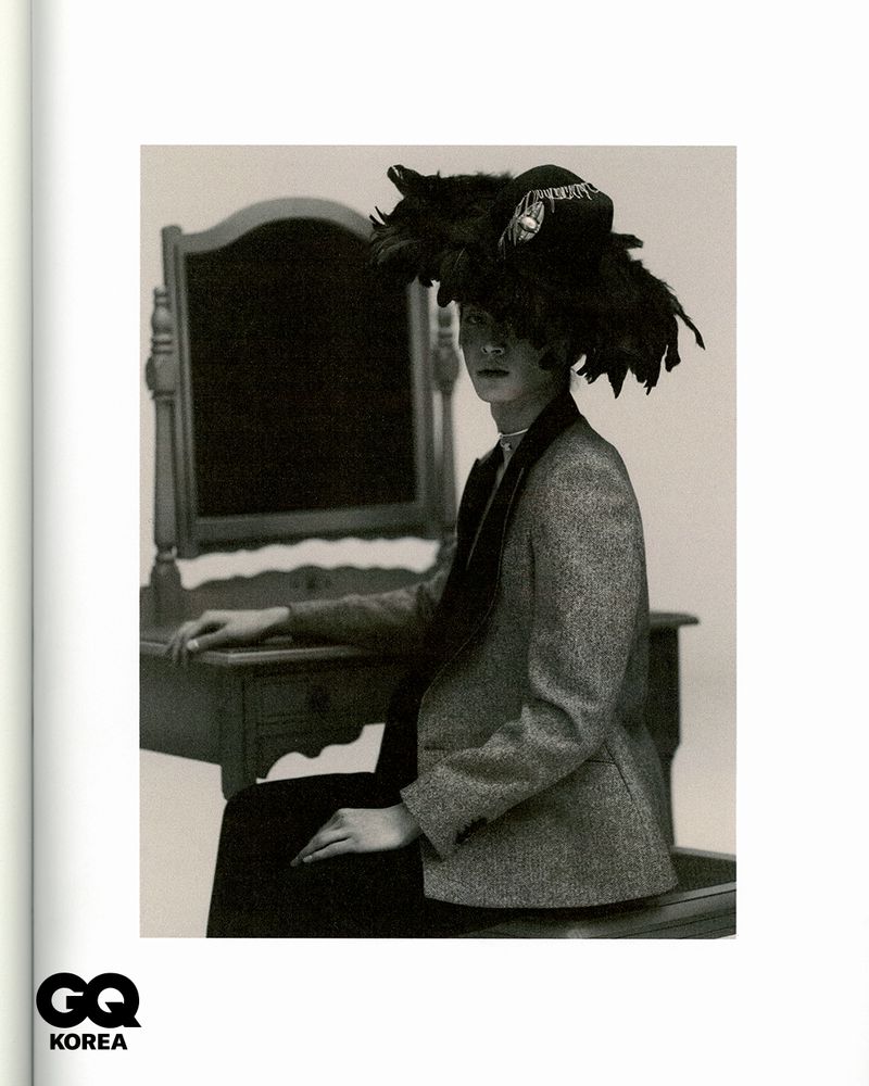 A seated person in a feathered hat and blazer poses in front of a vanity in a black and white fashion shot with GQ Korea logo.