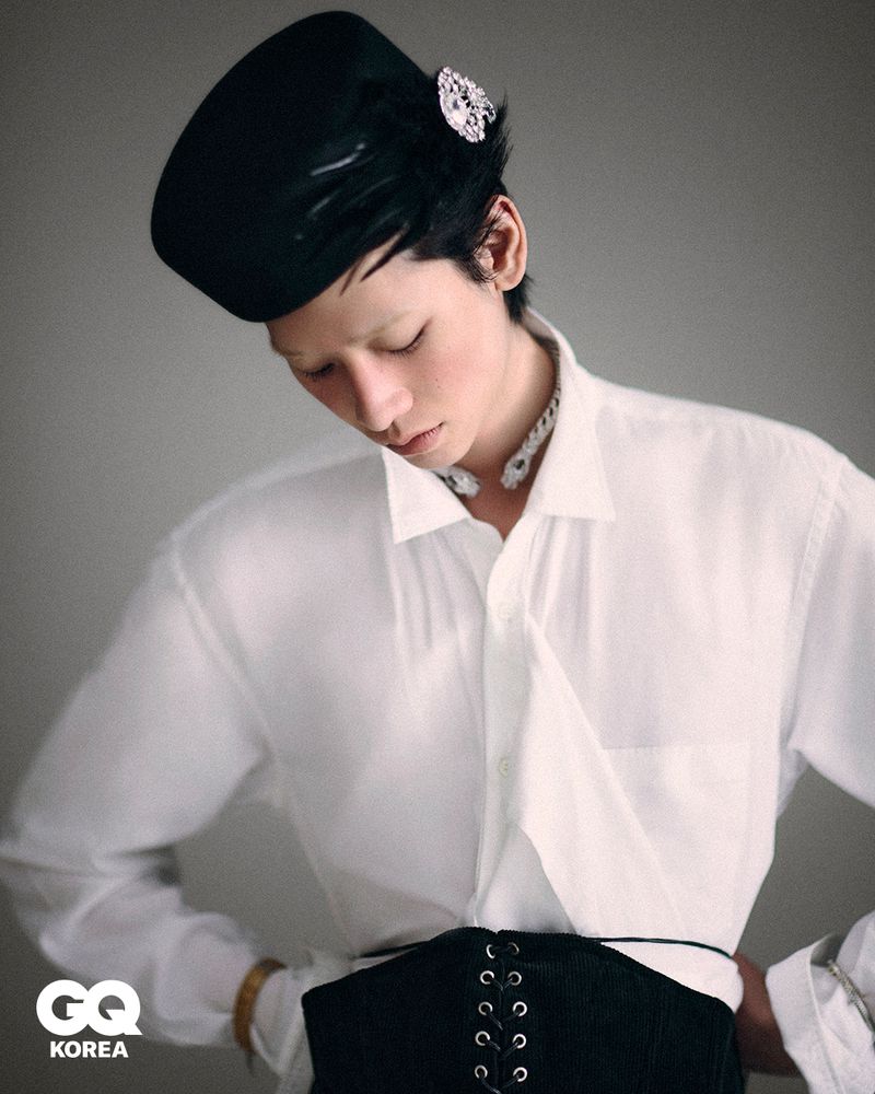 Model Yugo Takano wearing a white button-down shirt, black lace-up corset belt, and jeweled black beret against a grey background.