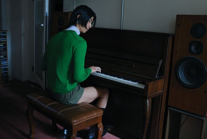 Model Yugo Takano seated at a dark upright piano playing keys in a green sweater beside large wooden speakers.