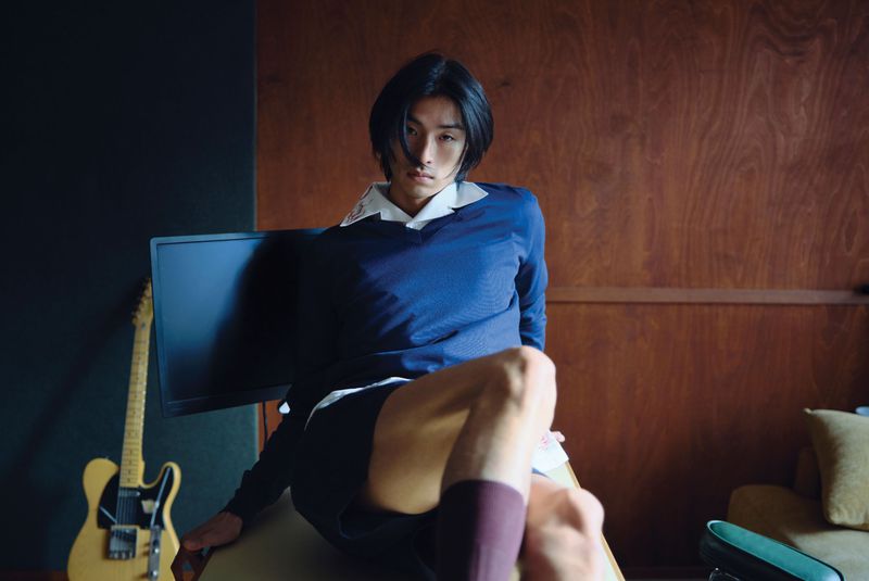 Young Asian man poses indoors with a serious expression in front of a guitar, monitor, wood wall and black wall wearing athletic clothing.
