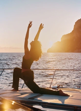 Model doing yoga pose on yacht deck at sunset with ocean and coastal cliffs in background for Alo Summer Campaign '26