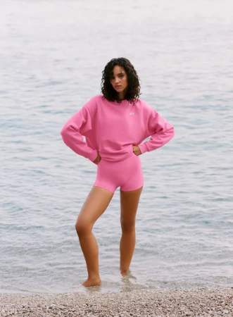 Model standing on a pebble shoreline wearing a bright pink Alo sweatshirt and matching shorts against coastal water.