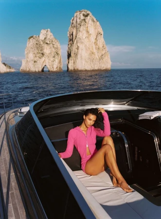 Model Rae Rodriguez in a bright pink zip-up hooded set reclining on a boat lounge with coastal sea stacks in the background.
