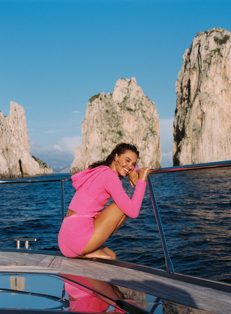 Smiling model in bright pink hoodie squatting on yacht deck with dramatic rock formations rising from blue water in background