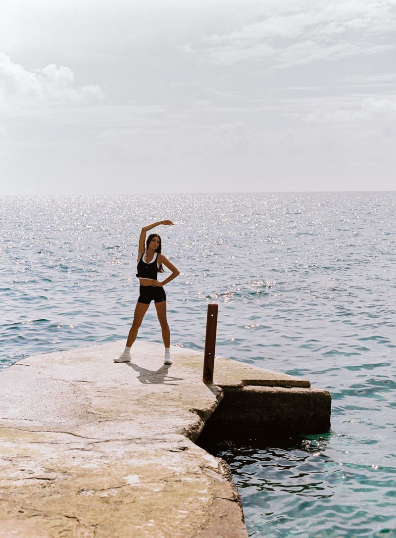 Model posing in black activewear and white sneakers with one arm raised on a concrete pier overlooking open blue water.