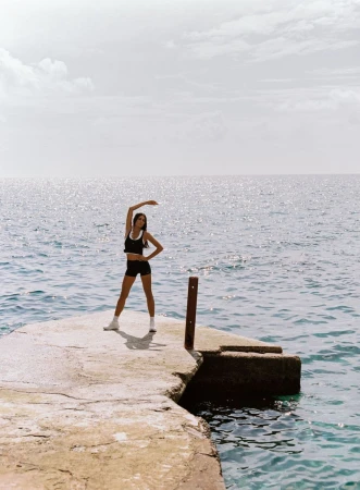 Model posing in black activewear and white sneakers with one arm raised on a concrete pier overlooking open blue water.