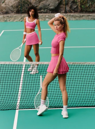 Frida Aasen and Rae Rodriguez wearing pink tennis sets with white sneakers on a green court for Alo.