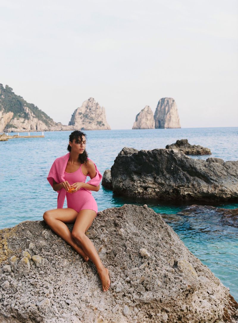 Model wearing a pink swimsuit seated on a coastal rock formation with ocean water and distant sea stacks behind her.