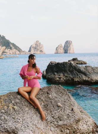 Model wearing a pink swimsuit seated on a coastal rock formation with ocean water and distant sea stacks behind her.