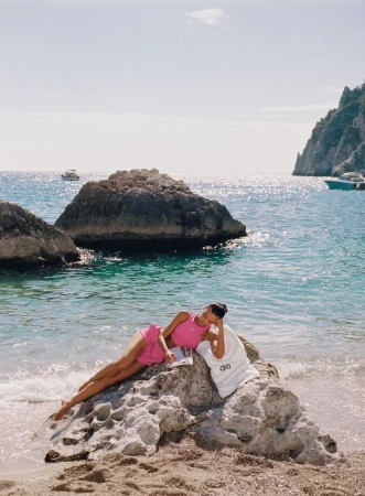 Woman in pink loungewear relaxes on a rock at the beach with a book and Alo brand tote bag, featuring boats in the distance.
