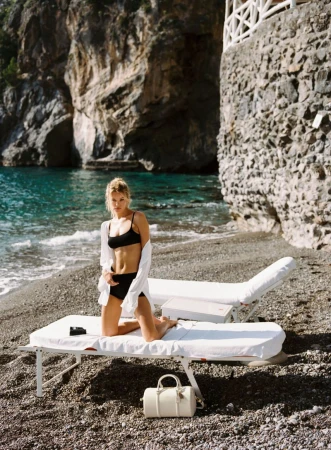 Model Frida Aasen kneeling on a white sun lounger in a black bikini and open white shirt on a pebbled rocky beach.