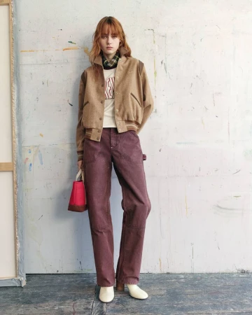 Model standing against a distressed wall wearing a camel jacket, plum trousers, and holding a small pink and brown handbag.