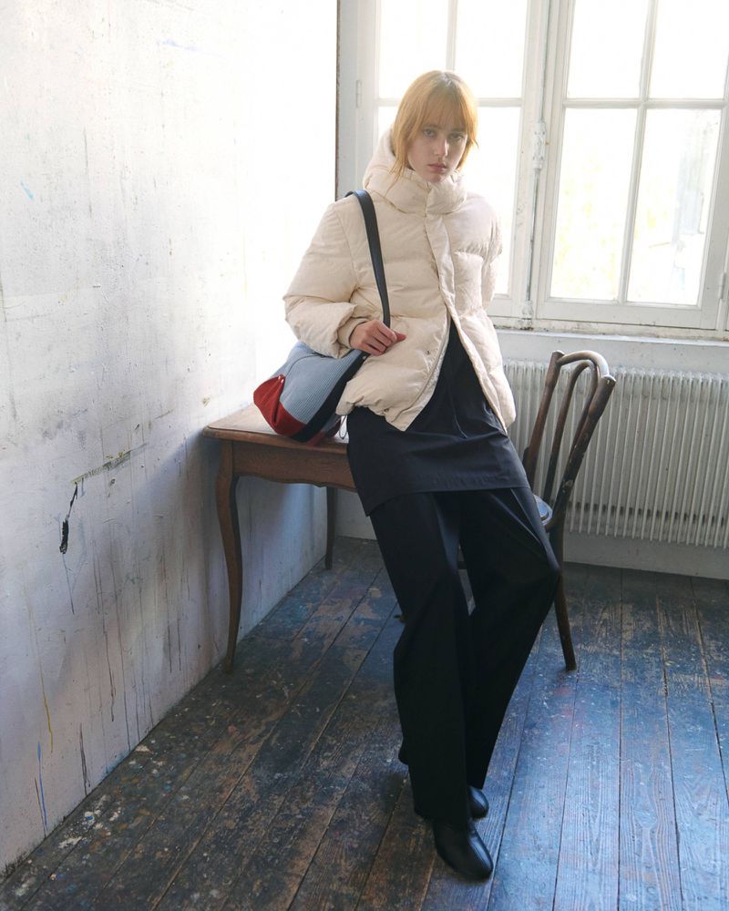 Model leaning against a wooden table wearing a cream puffer jacket, wide-leg black trousers, and a blue shoulder bag.