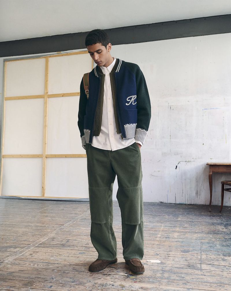 Model standing in a studio wearing a blue and green varsity jacket, white shirt, and green trousers.