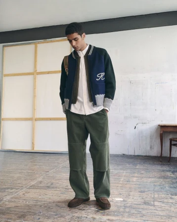 Model standing in a studio wearing a blue and green varsity jacket, white shirt, and green trousers.