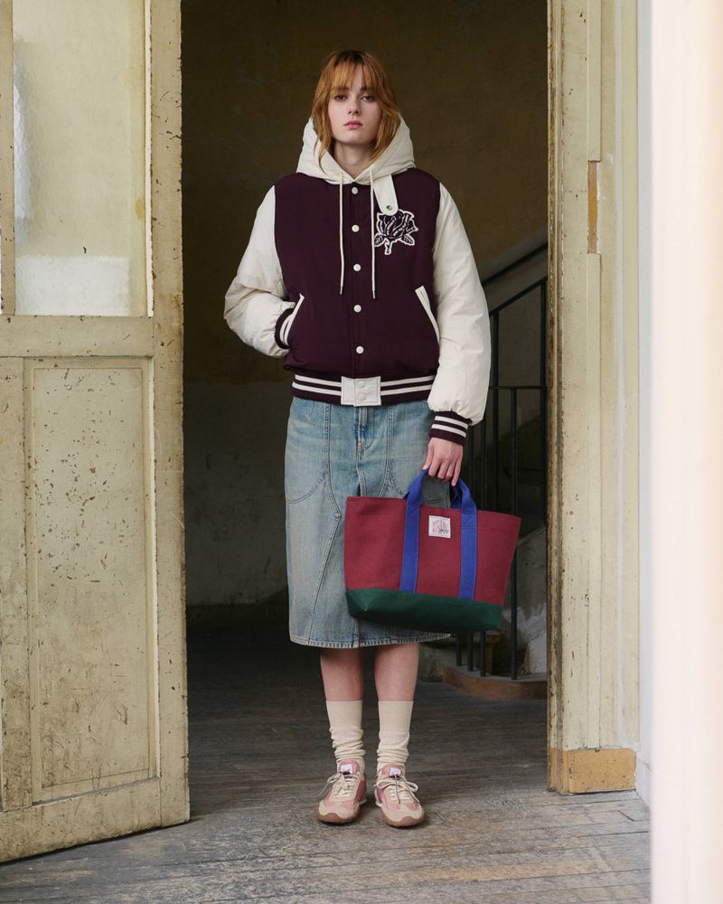 Model in a hooded burgundy varsity jacket and denim midi skirt holding a colorblocked tote bag in an open doorway.