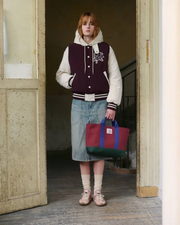 Model in a hooded burgundy varsity jacket and denim midi skirt holding a colorblocked tote bag in an open doorway.