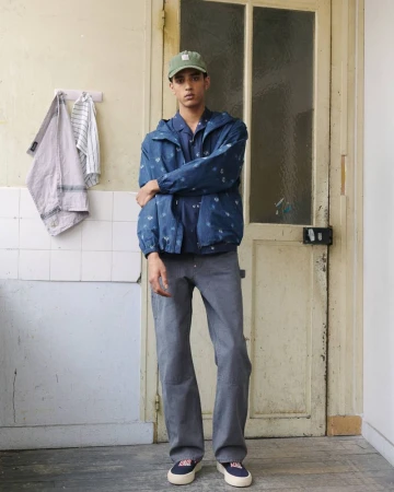 Model wearing a blue patterned jacket and grey trousers standing indoors against an old wooden door and tiled wall.