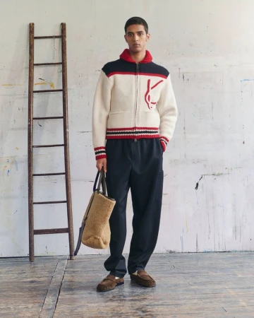 Model standing in a white zip-up knit sweater, dark trousers, and holding a fleece tote bag next to a rustic wooden ladder.