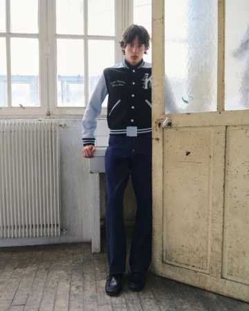 Model wearing a varsity jacket and dark trousers standing indoors near a doorway and white radiator
