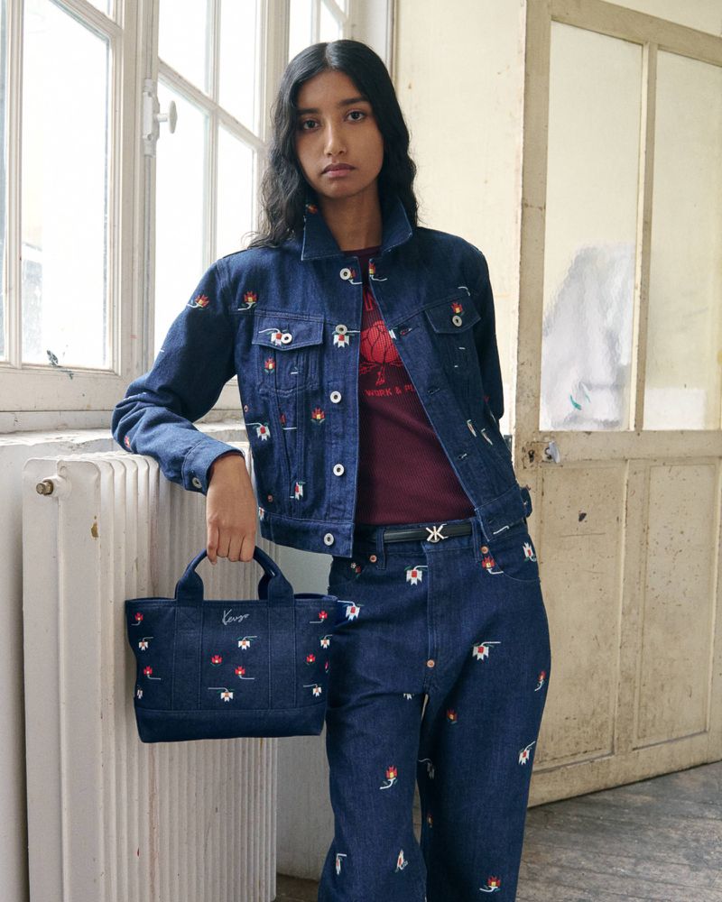 Model wearing embroidered denim jacket and matching jeans holding a blue denim handbag next to a radiator.