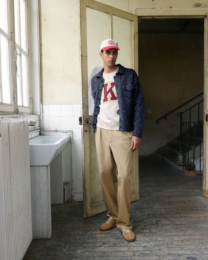 Male model wearing a textured navy jacket, varsity letter sweatshirt, beige trousers, and a baseball cap standing in an aged interior.