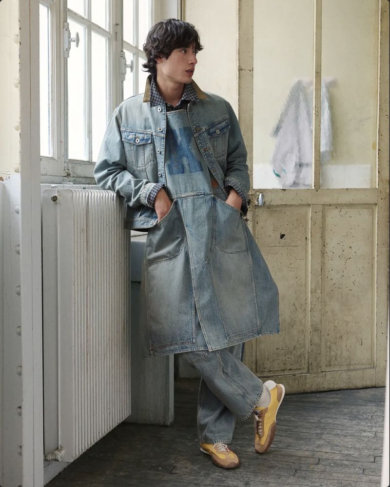 A young male model wearing layered denim apparel leans against a radiator near a large window in a room.