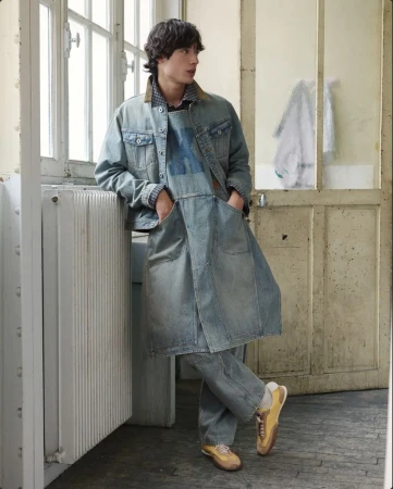A young male model wearing layered denim apparel leans against a radiator near a large window in a room.
