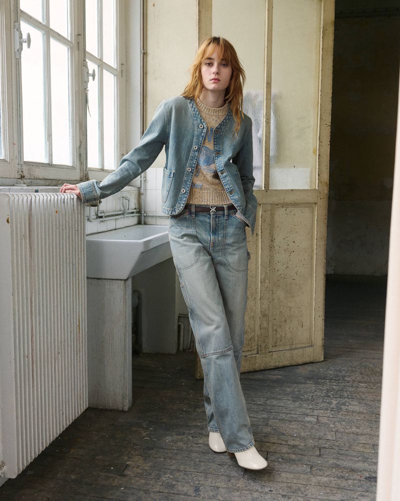 Model standing by a large window wearing a light wash denim jacket with matching wide-leg jeans, patterned knit top, and white boots.