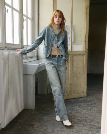 Model standing by a large window wearing a light wash denim jacket with matching wide-leg jeans, patterned knit top, and white boots.