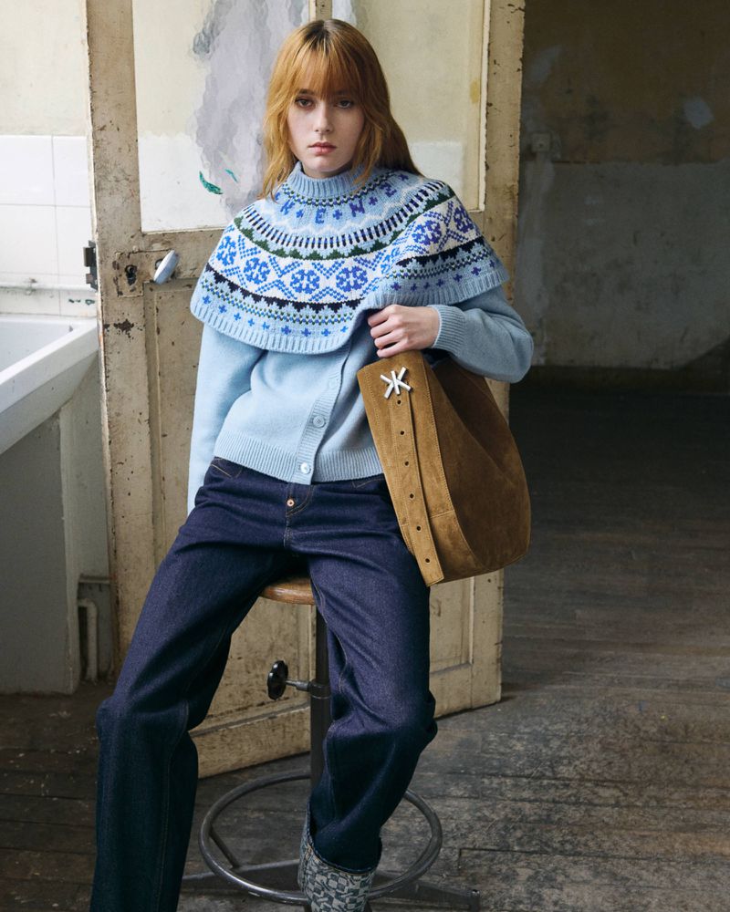 Model seated on a stool wearing a blue patterned knit sweater and dark denim jeans holding a brown suede bag