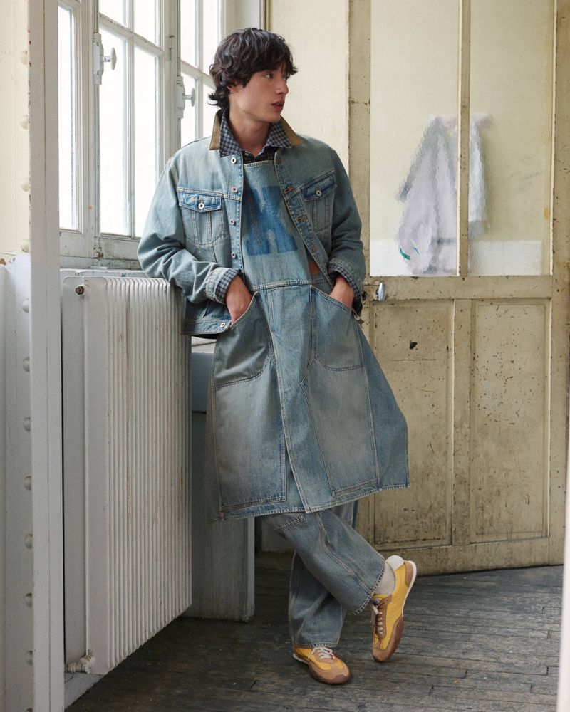A male model leaning against a white radiator wearing a layered denim jacket, long apron tunic, matching jeans, and yellow sneakers.