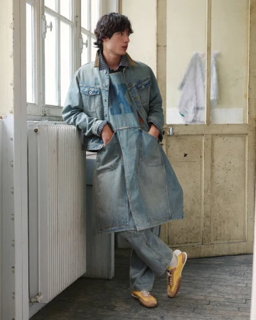 A male model leaning against a white radiator wearing a layered denim jacket, long apron tunic, matching jeans, and yellow sneakers.